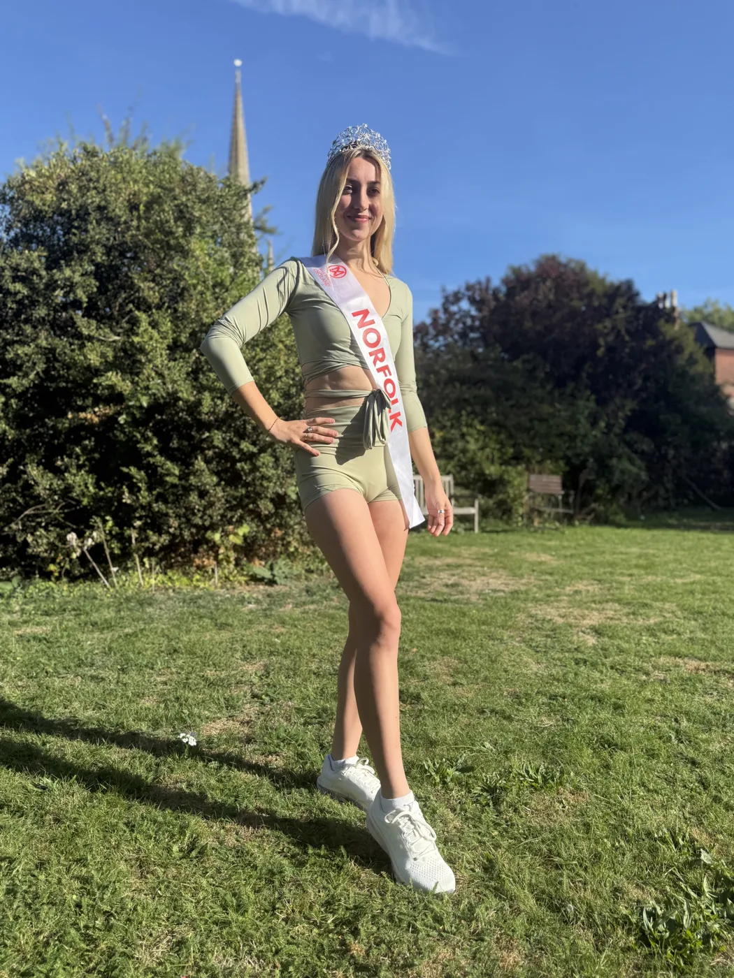 Amy Morris standing on a lawn in sportswear, trainers, a crown and a Norfolk sash, with trees and a spire behind